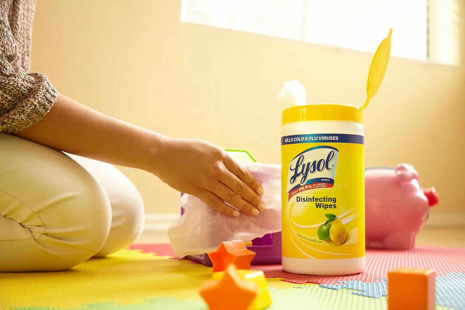Parent kneeling on floor and wiping baby toys next to canister of Lysol Disinfectant wipes