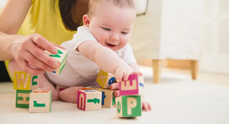 Parent helps baby build stack of lettered blocks when sitting on floor