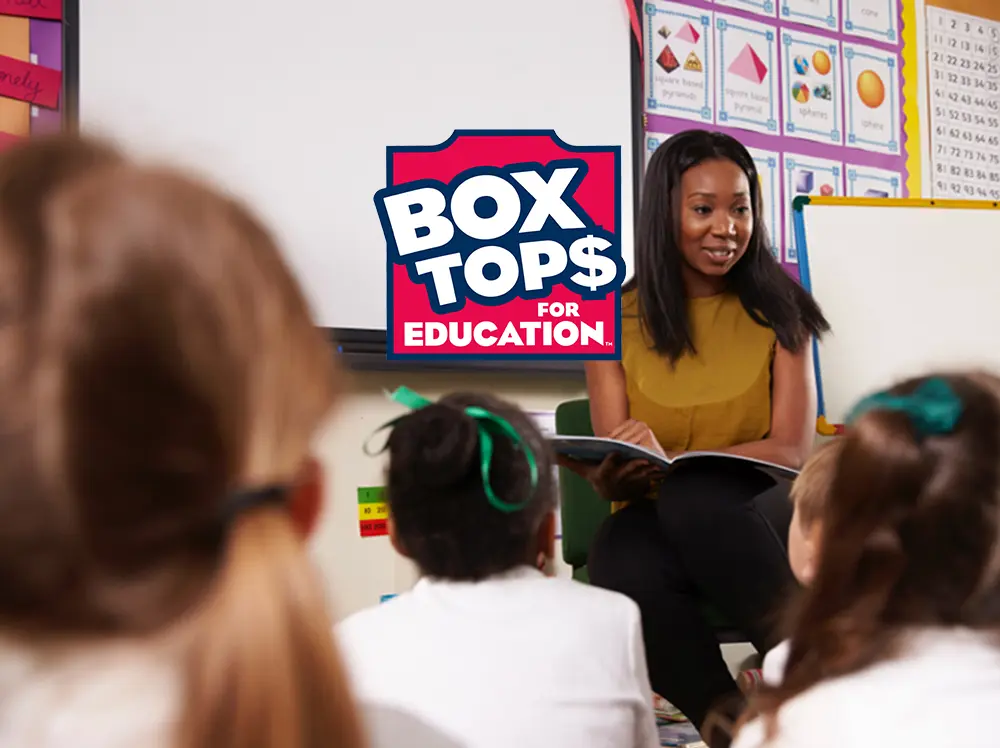 A teacher at the front of a classroom reading a book to a group of elementary students. Box Tops for Education Logo overlays the image.