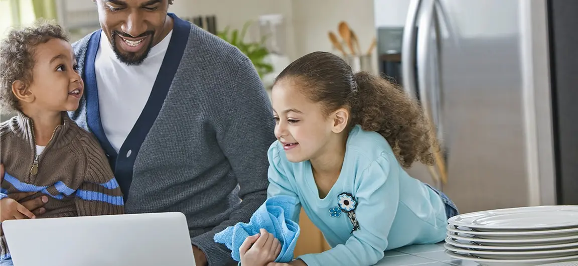 A parent and two children in a kitchen. The parent is showing the children something on a laptop computer. One of the children has a drying cloth in their hand and is next to clean and dry dishes.