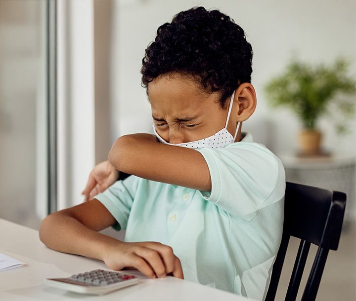 A child wearing a mask sneezes into their arm while sitting at a table with a calculator.