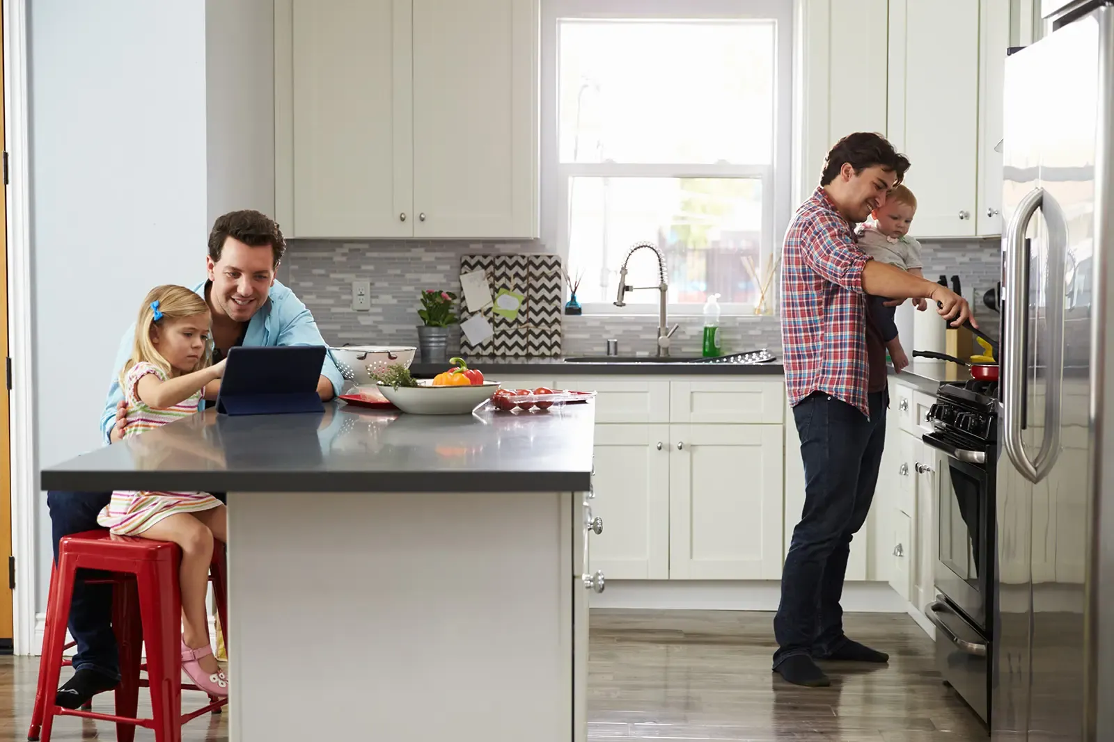 One parent and child sat at kitchen counter looking at screen and another parent holds child while cooking on stove